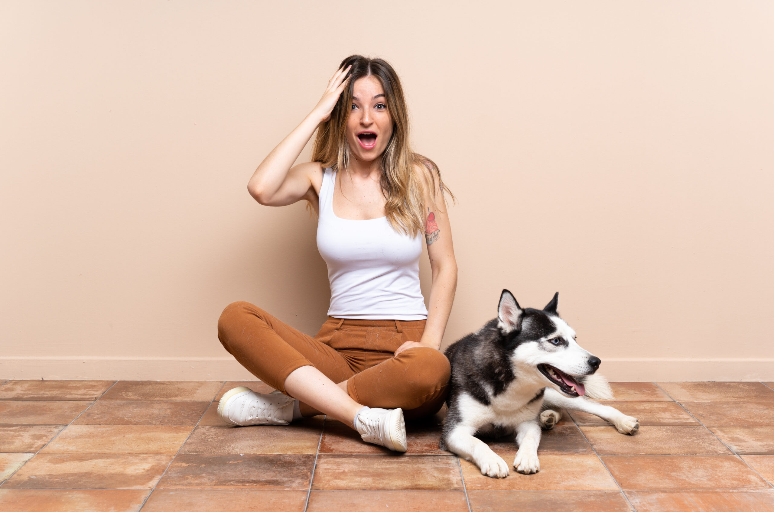 Young pretty woman with her husky dog sitting in the floor at indoors with surprise and shocked facial expression