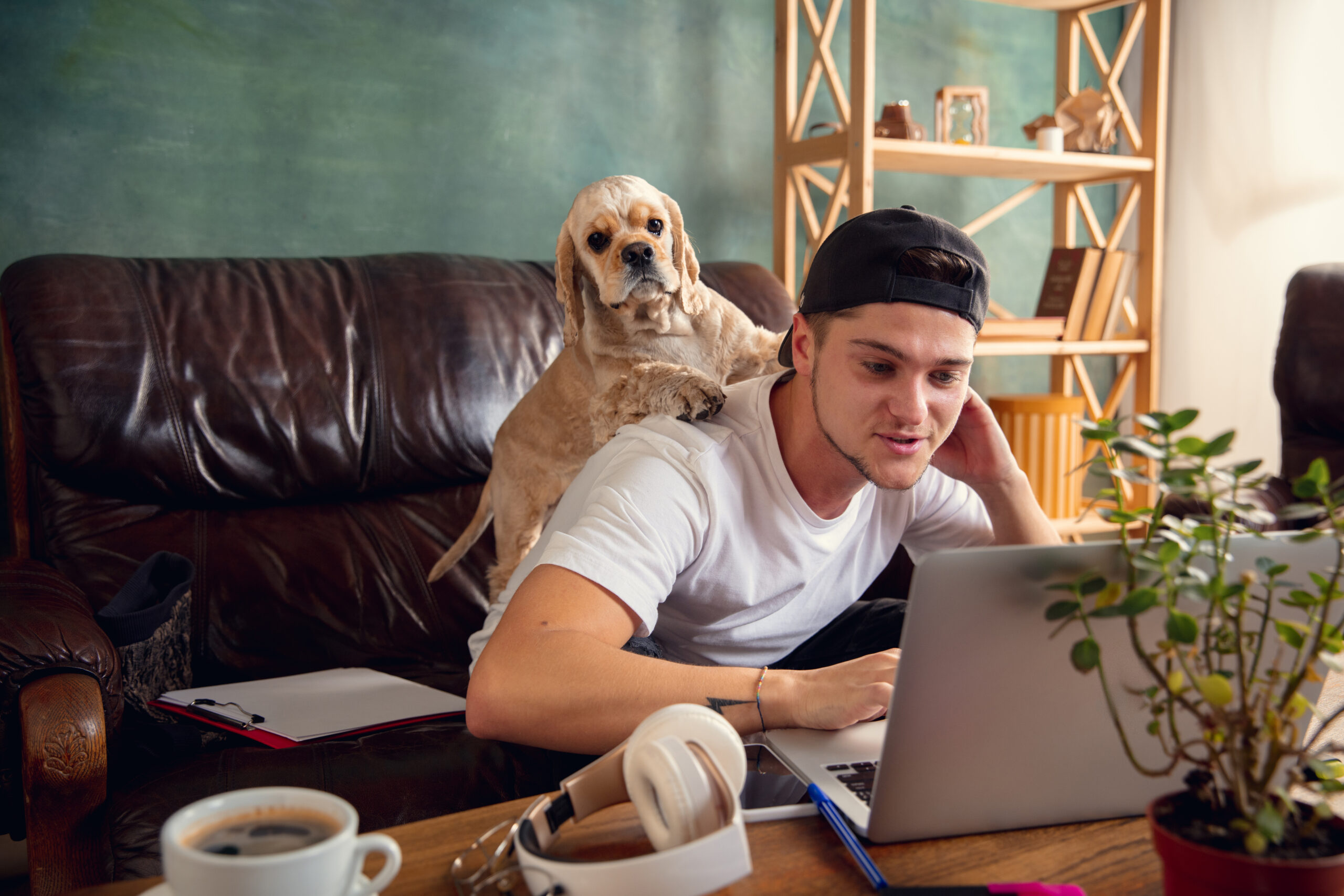 Young handsome man sitting and working at home with his cute dog. Cozy office workplace, remote work, online learning concept.